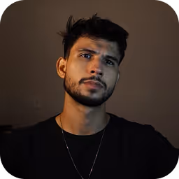 Portrait of a young man with dark hair and beard wearing a black shirt and a thin chain necklace against a dark background.